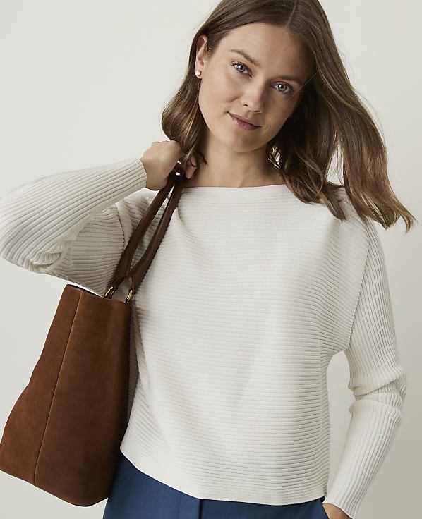 A woman in a white ribbed sweater and blue pants carries a brown tote bag against a plain background.