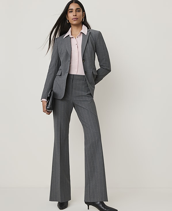 A woman in a gray pinstripe suit and pink blouse stands against a plain white background, holding a black clutch.