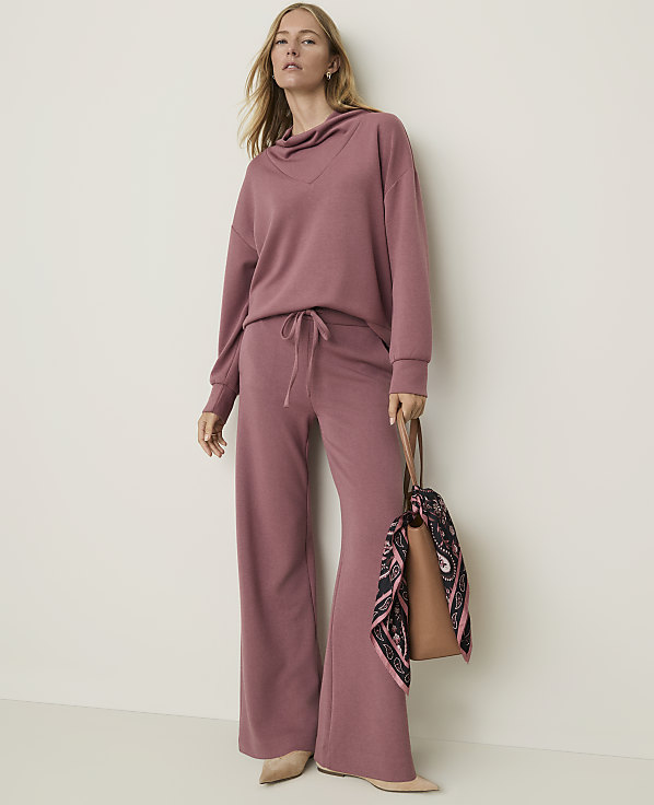 A woman in a mauve loungewear set stands against a plain, light-colored wall, holding a brown tote bag with a patterned scarf.