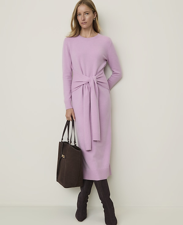 A woman in a light purple dress and dark boots stands with a brown bag against a plain white background.
