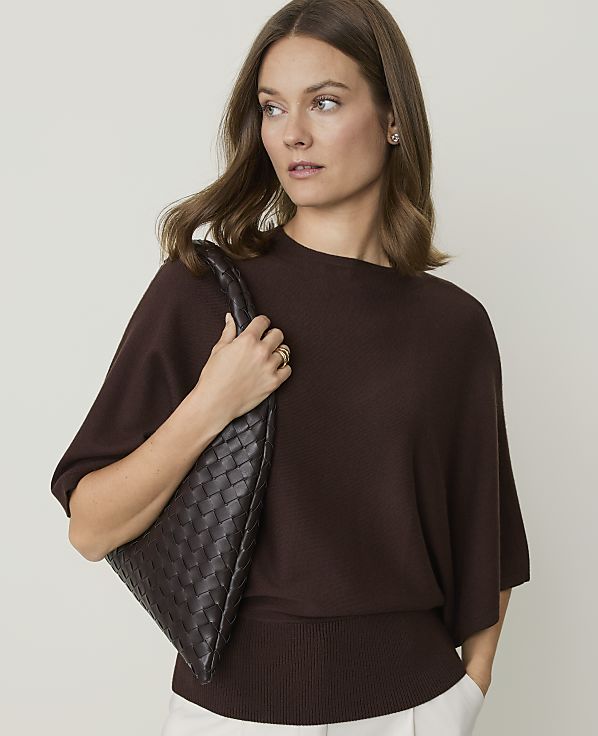 A woman in a brown sweater and white pants holds a woven brown purse against a plain background.