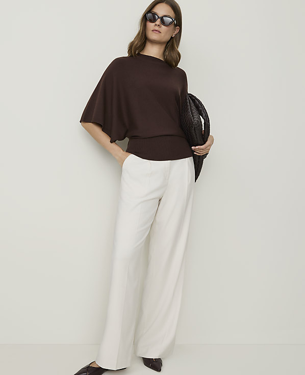 A woman in a brown sweater and white pants stands against a plain background, holding a woven bag.