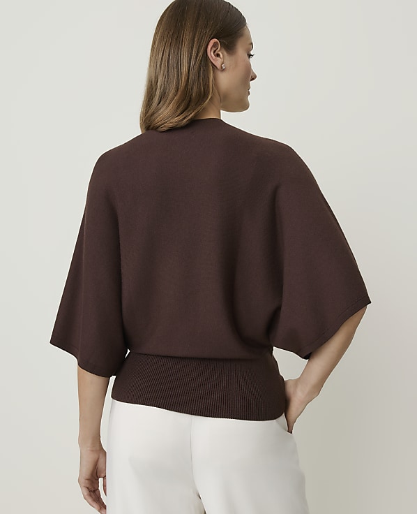A woman in a brown sweater and white pants stands against a plain white background.