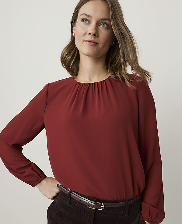 A woman in a rust-colored blouse and dark pants stands against a plain, light background.