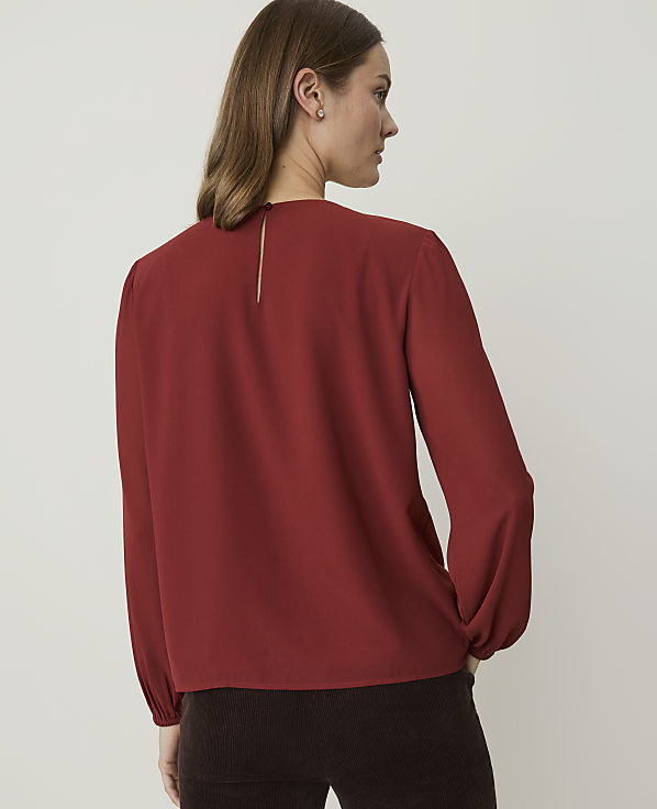 A woman in a rust-colored blouse stands against a plain, light background.