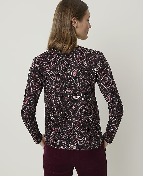 A woman in a black paisley-patterned shirt stands with her hands in her pockets against a plain, light-colored background.