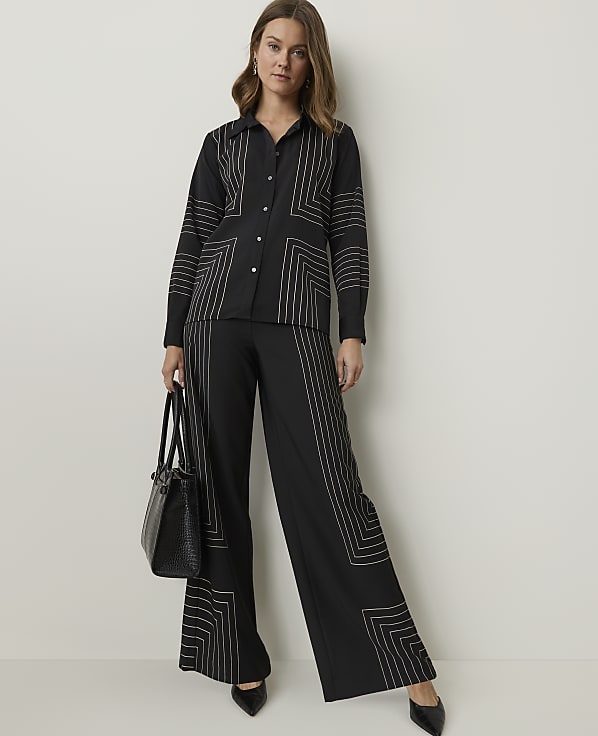 A woman in a black patterned shirt and wide-leg pants stands against a plain white background, holding a black handbag.