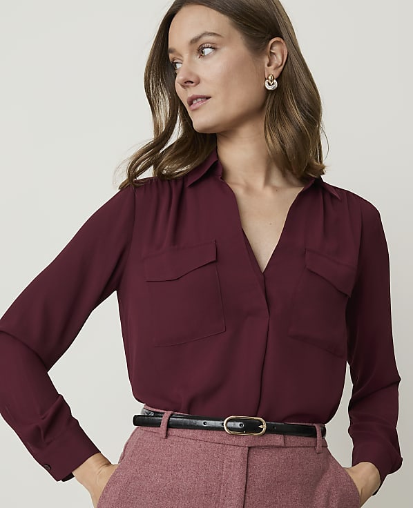 A woman in a burgundy blouse and pink pants stands against a plain, light background.