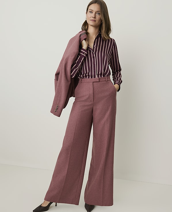 A woman in a striped blouse and wide-leg pants stands against a plain, light-colored background, with a blazer draped over her shoulder.