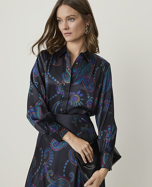 A woman in a dark, paisley-patterned blouse and skirt holds a black clutch against a plain, light background.
