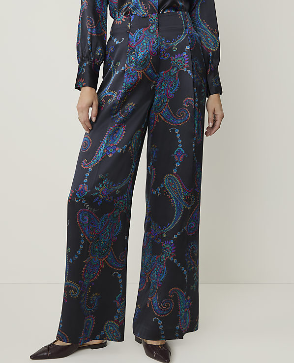 A person stands in wide-leg, dark satin pants with a colorful paisley pattern, against a plain, light-colored background.