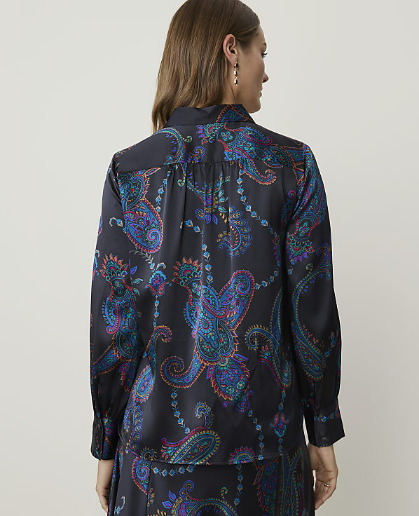A woman in a dark, patterned blouse stands against a plain background.