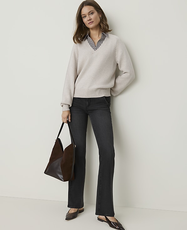 A woman in a cream v-neck sweater and dark wash jeans stands against a plain, light-colored wall, holding a brown and black patchwork bag.