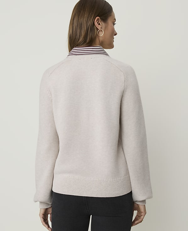 A woman in a light-colored sweater and dark pants stands against a plain white background.