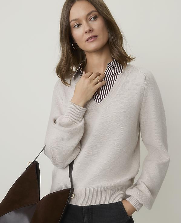 A woman in a cream v-neck sweater over a striped shirt stands against a plain, light background, with a brown bag in the foreground.