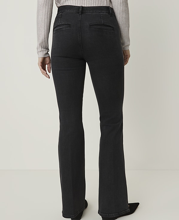 A person wearing dark wash flared jeans and a light grey sweater stands against a plain white background.