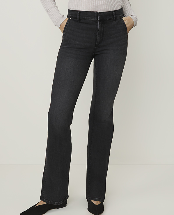 A person stands in dark wash flared jeans and black pointed-toe flats against a plain, light gray background.