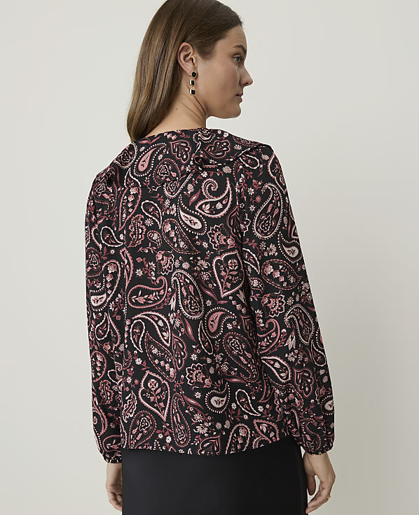 A woman in a black paisley-patterned blouse stands against a plain white background.