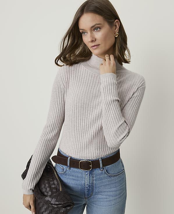 A woman in a beige turtleneck and jeans holds a dark woven bag in front of a plain background.