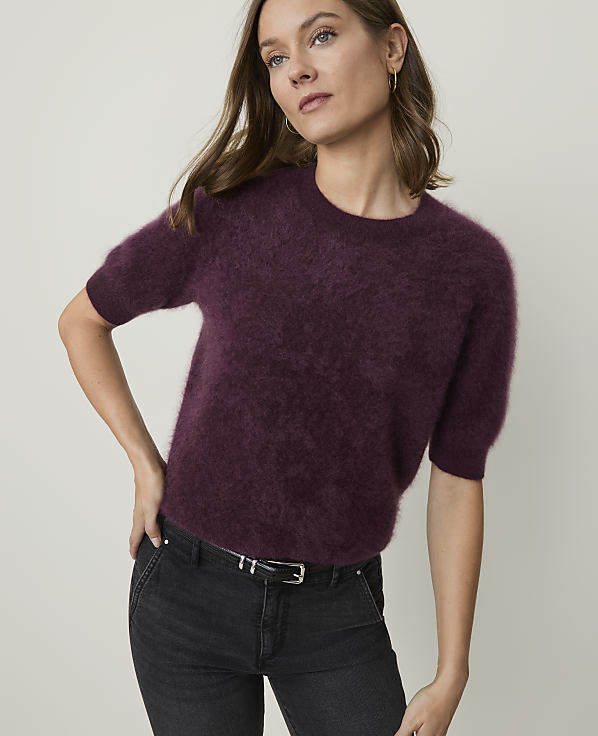 A woman in a fuzzy plum-colored sweater and dark jeans stands against a plain, light gray background.