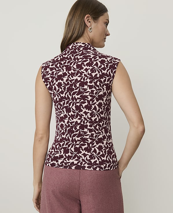 A woman in a floral top and pink pants stands against a plain background.