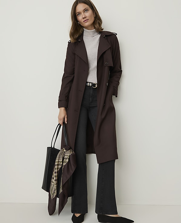 A woman in a dark brown trench coat and black flared jeans stands against a plain white background, holding a black tote bag with a patterned scarf.