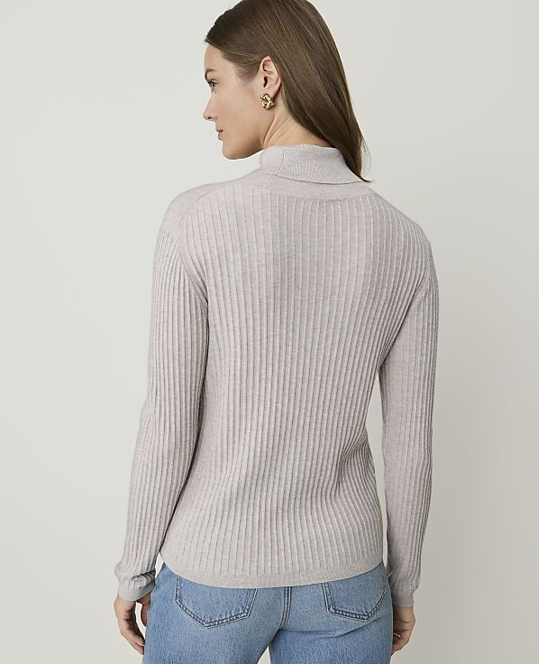 A woman in a light gray ribbed turtleneck sweater and blue jeans stands against a plain, light gray background.
