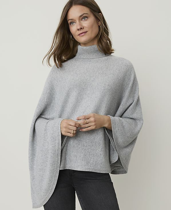 A woman in a grey turtleneck poncho stands against a plain grey background.