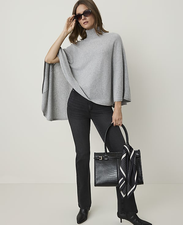 A woman in a gray poncho and black jeans poses with a black handbag against a plain gray background.