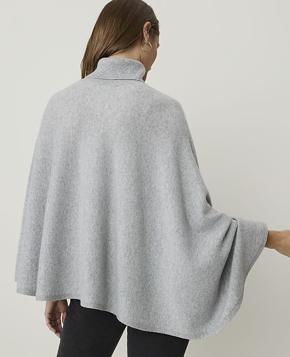 A woman wearing a light gray turtleneck poncho stands against a plain, light gray background.
