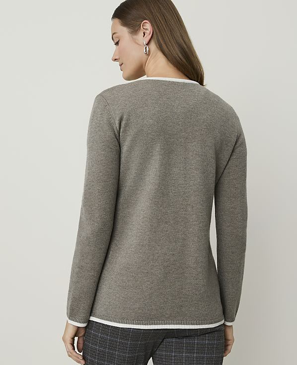 A woman in a taupe sweater with white trim stands against a plain, light gray background.