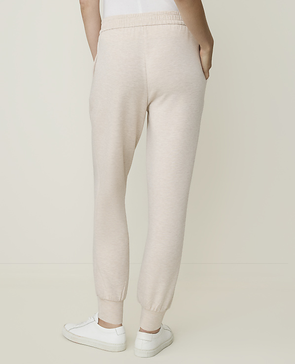 A person wearing light-colored sweatpants and white sneakers stands against a plain, light-colored background.