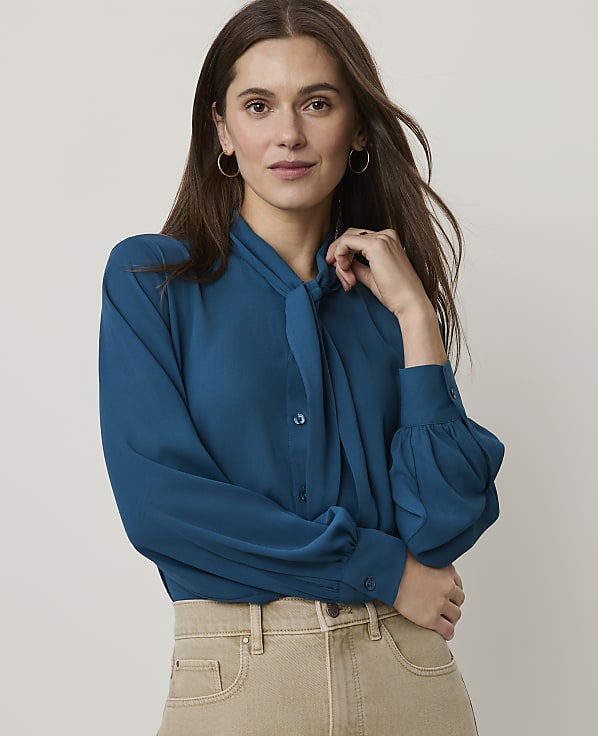 A woman in a teal blouse and tan pants poses against a plain white background.