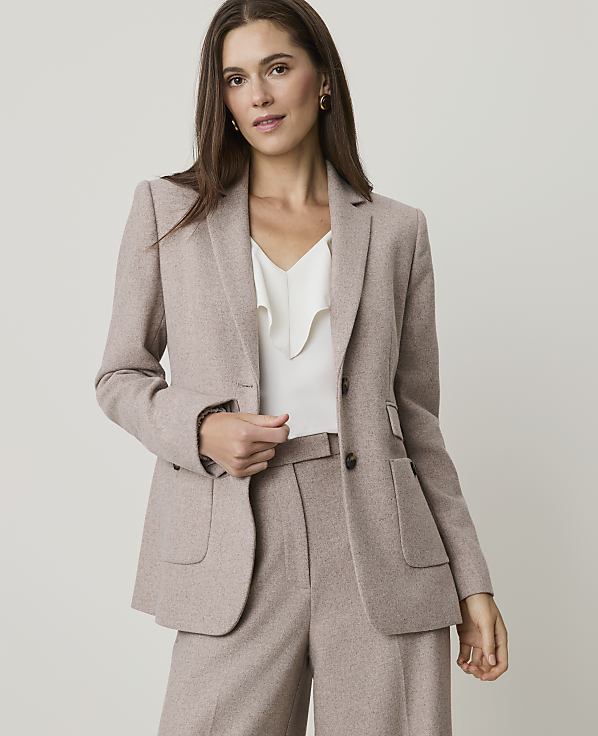 A woman in a beige blazer and pants stands against a plain gray background.