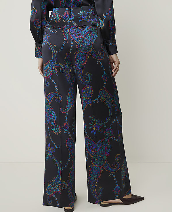 A person stands in wide-leg paisley-patterned pants and dark flats in the foreground, against a plain, light-colored background.