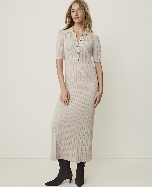 A woman in a beige cable-knit midi dress and dark boots stands against a plain, light-colored background.