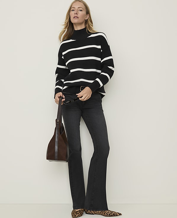 A woman in a black and white striped turtleneck sweater and dark flared jeans stands against a plain white background, holding a brown leather handbag and wearing leopard print flats.