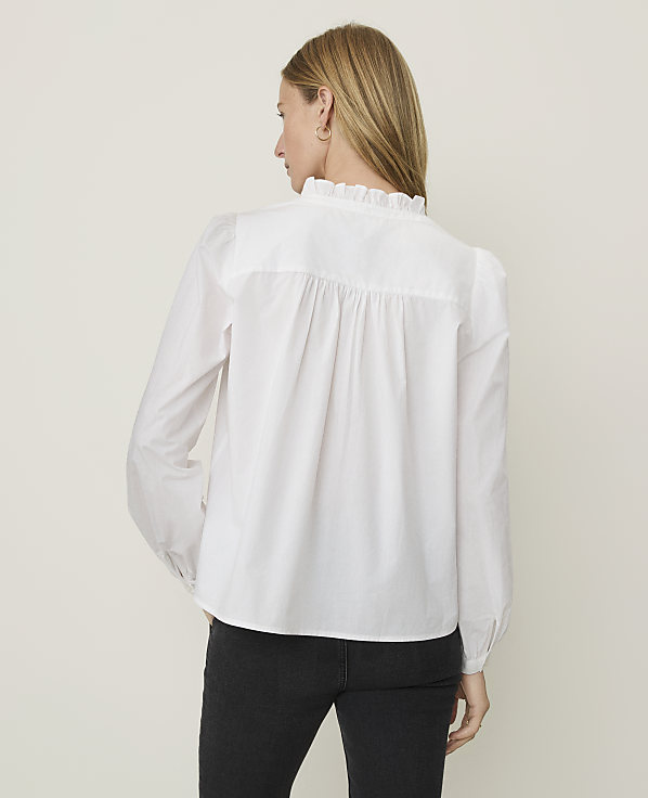 A woman in a white ruffled blouse stands against a plain, light-colored background.