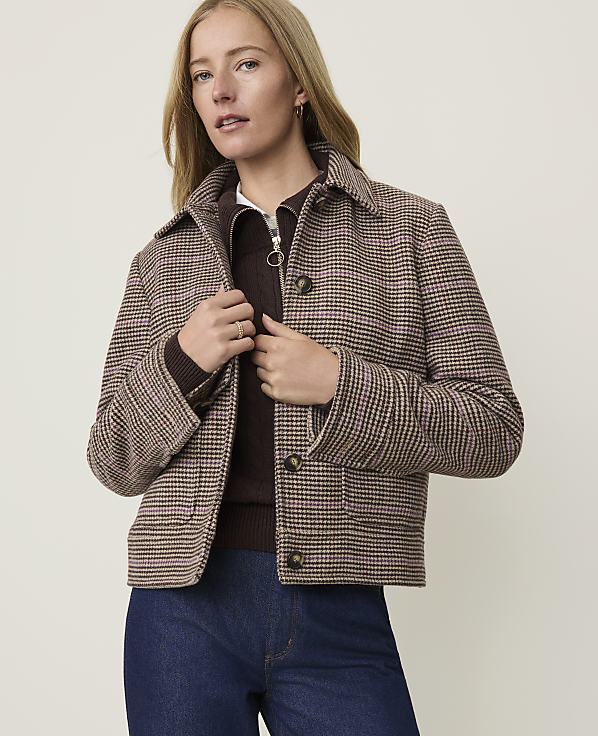 A woman in a plaid jacket and dark jeans stands against a plain, light-colored background.