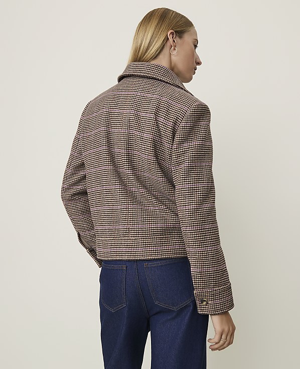 A woman in a brown and pink plaid jacket and dark jeans stands against a plain, light-colored background.