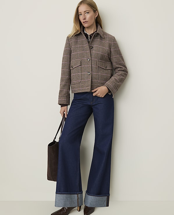 A woman in a plaid jacket and wide-leg jeans stands against a plain, light-colored wall, holding a brown tote bag.