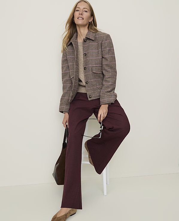 A woman in a patterned jacket and maroon wide-leg pants sits on a stool with a brown bag, against a plain, light-colored background.