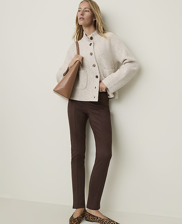 A woman in a cream jacket and brown pants stands against a plain background, carrying a brown purse.