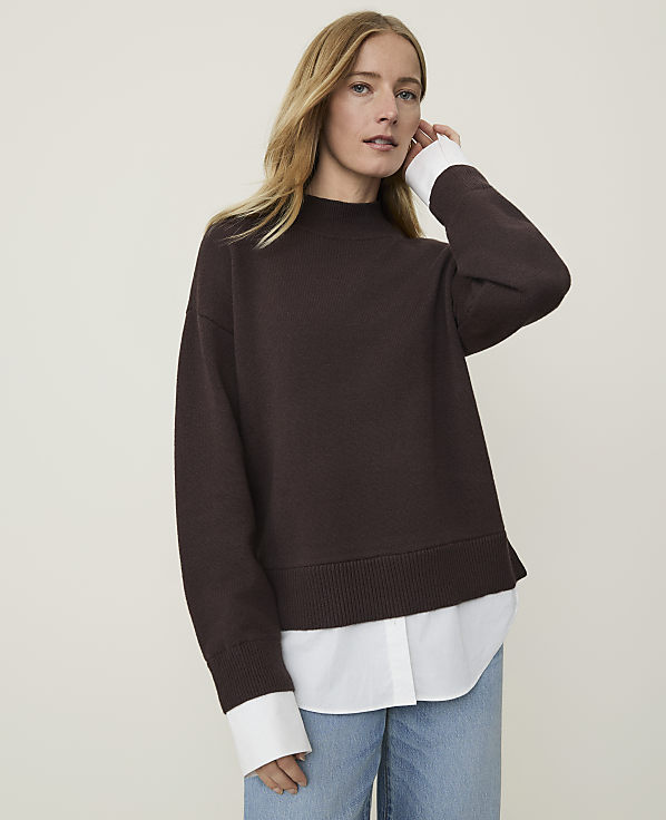 A woman in a brown sweater layered over a white shirt stands against a plain, light-colored background.