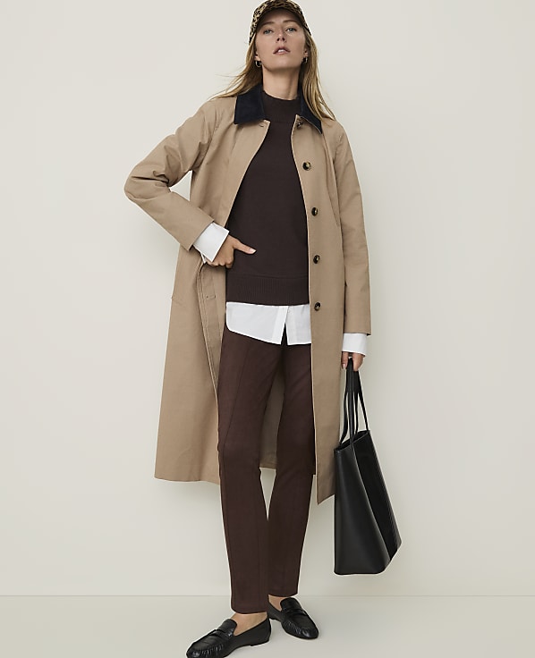A woman in a trench coat and brown pants stands against a plain, light-colored background, holding a black tote bag.