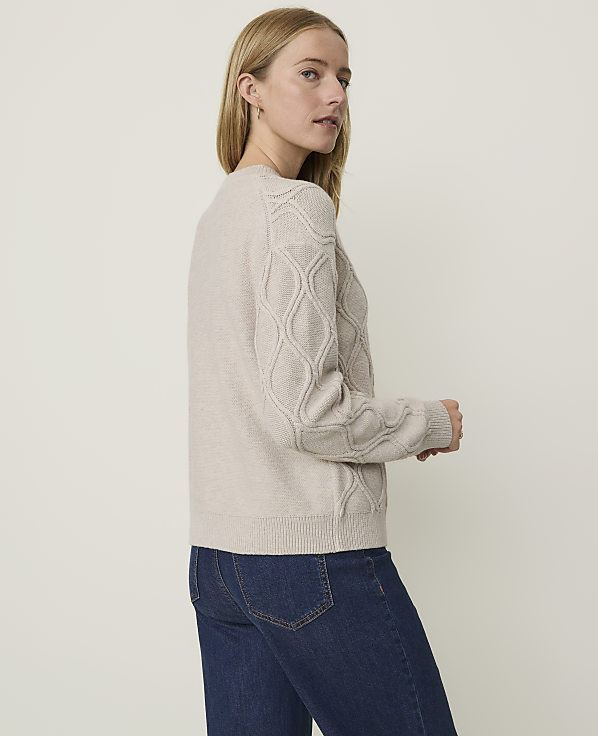 A woman in a beige cable-knit sweater and blue jeans stands against a plain, light-colored background.