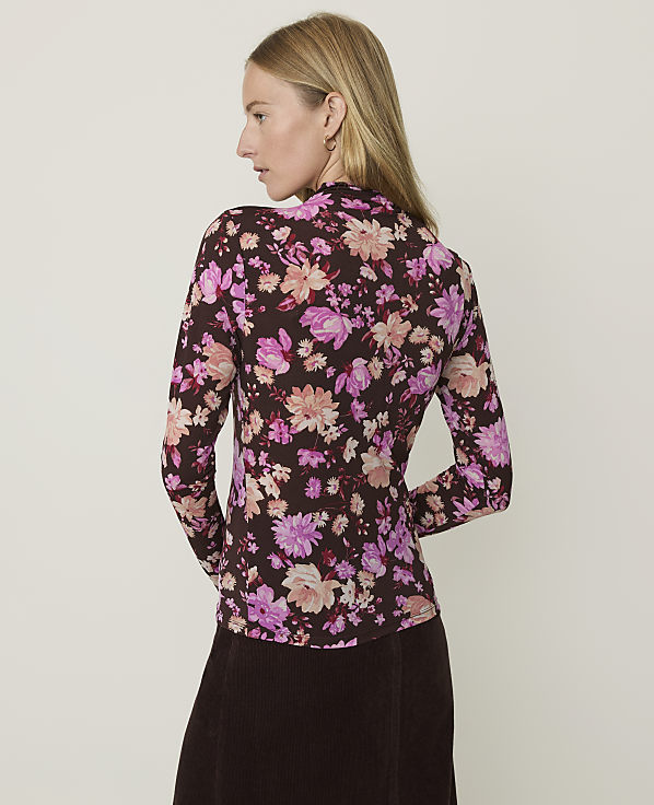 A woman in a floral top and brown skirt stands against a plain, light background.
