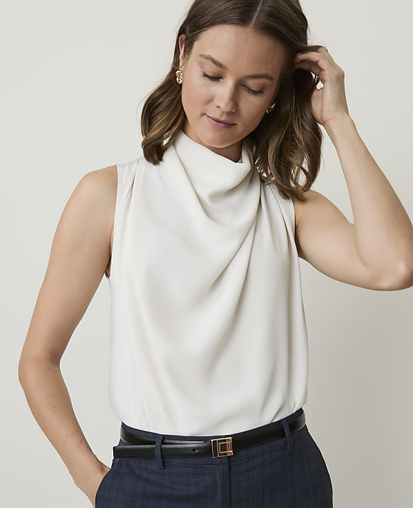 A woman in a cream cowl-neck sleeveless top and dark pants stands against a plain, light background.