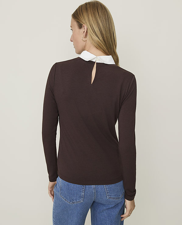 A woman in a brown long-sleeved shirt with a white collar and blue jeans stands against a plain beige background.