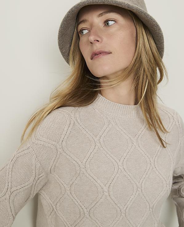 A woman in a beige sweater and hat looks to the side against a plain background.